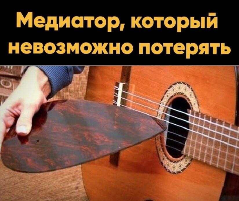 Рука держит гигантский деревянный медиатор, который невозможно потерять, рядом с гитарой