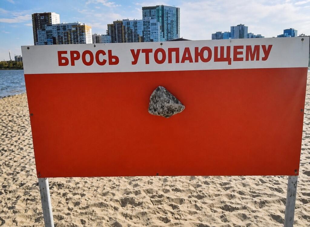 Красно-белый спасательный стенд на пляже с надписью БРОСЬ УТОПАЮЩЕМУ, на месте спасательного круга прикреплён серый камень